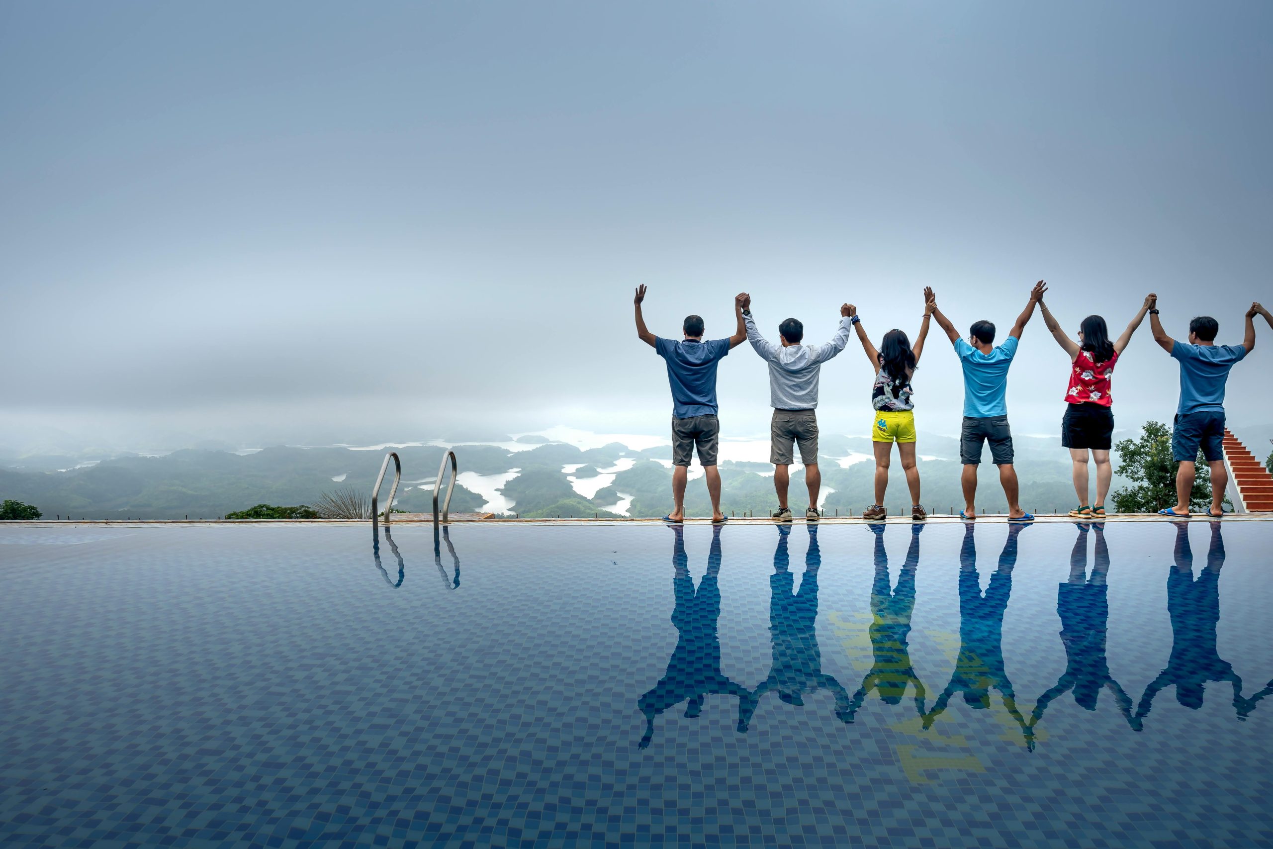 Seven people standing by an infinity pool with arms raised, reflected in water, overlooking scenic foggy landscape.